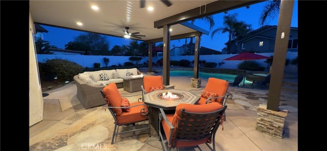 patio at night with a fenced backyard, ceiling fan, an outdoor living space with a fire pit, and a fenced in pool