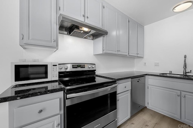 kitchen featuring a sink, dark stone counters, under cabinet range hood, light wood finished floors, and appliances with stainless steel finishes
