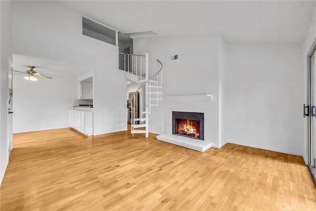 unfurnished living room featuring a ceiling fan, stairs, a brick fireplace, a towering ceiling, and light wood-style floors