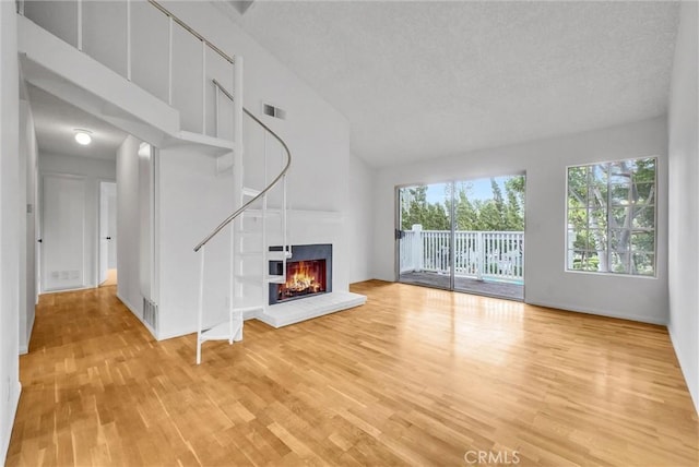 unfurnished living room featuring high vaulted ceiling, a textured ceiling, light wood-type flooring, visible vents, and a glass covered fireplace