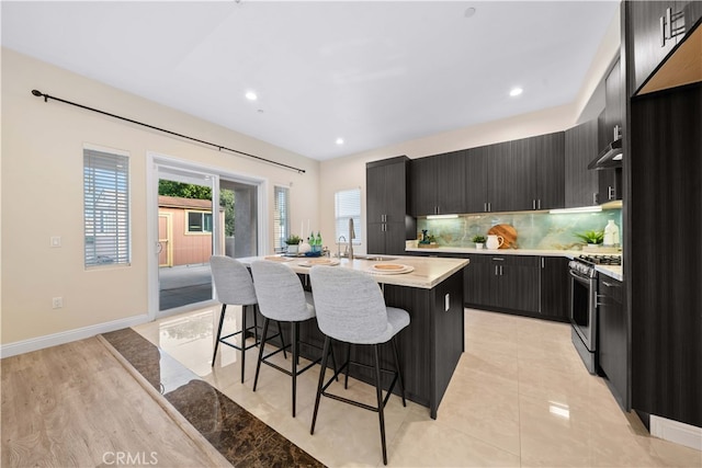 kitchen with a kitchen breakfast bar, a center island with sink, gas range, light countertops, and dark cabinetry