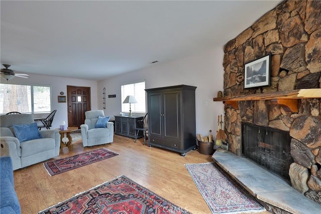 living room featuring a ceiling fan, a wealth of natural light, a stone fireplace, and light wood finished floors