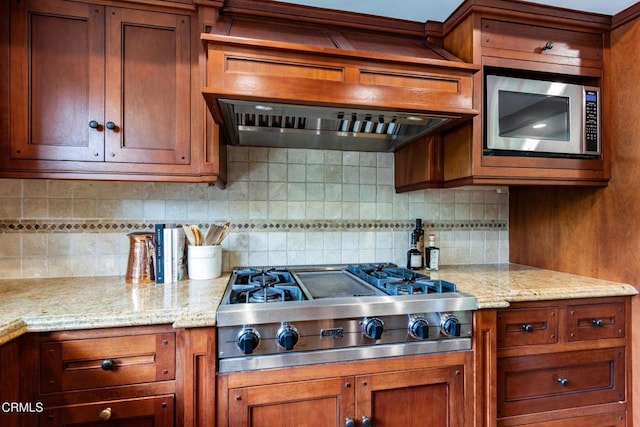 kitchen with light stone countertops, brown cabinetry, premium range hood, backsplash, and appliances with stainless steel finishes