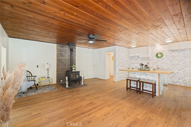 living room featuring light wood-style flooring, brick wall, ceiling fan, a wood stove, and wooden ceiling