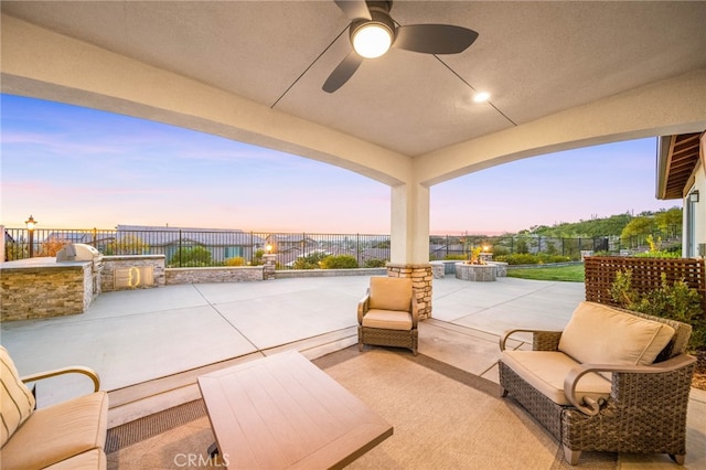 patio terrace at dusk featuring an outdoor kitchen, a fenced backyard, ceiling fan, and an outdoor fire pit