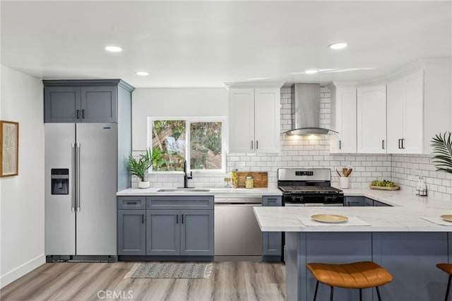 kitchen featuring light wood finished floors, a sink, a kitchen breakfast bar, wall chimney range hood, and stainless steel appliances