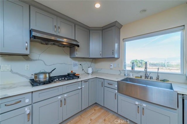 kitchen featuring light wood finished floors, a sink, under cabinet range hood, gray cabinetry, and stainless steel gas stovetop