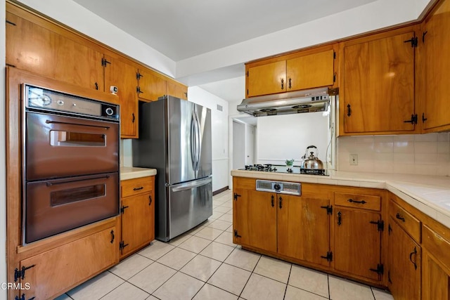 kitchen featuring tasteful backsplash, brown cabinets, freestanding refrigerator, under cabinet range hood, and multiple ovens
