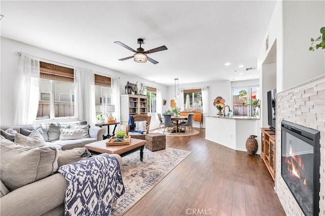 living area featuring a wealth of natural light, a glass covered fireplace, ceiling fan, and dark wood-style floors