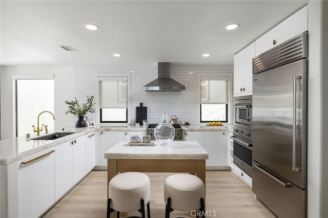 kitchen featuring a peninsula, a sink, wall chimney exhaust hood, white cabinetry, and appliances with stainless steel finishes