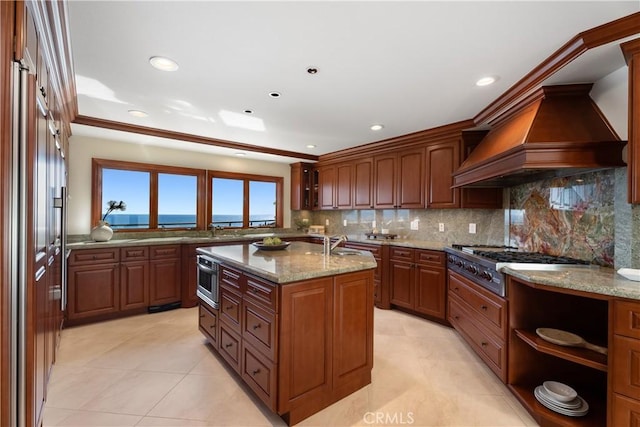 kitchen with stainless steel gas stovetop, a kitchen island with sink, light stone counters, backsplash, and light tile patterned floors