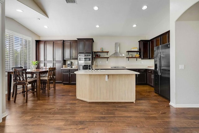 kitchen with open shelves, wall chimney exhaust hood, an island with sink, light countertops, and appliances with stainless steel finishes