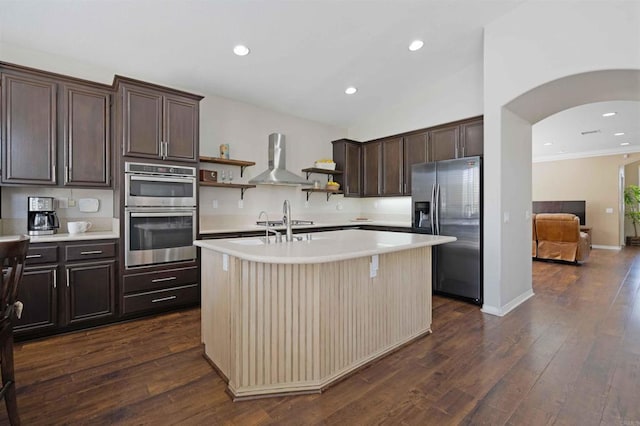 kitchen with wall chimney range hood, open shelves, light countertops, dark brown cabinets, and appliances with stainless steel finishes
