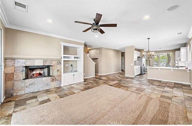 unfurnished living room featuring baseboards, ornamental molding, a tiled fireplace, visible vents, and ceiling fan with notable chandelier