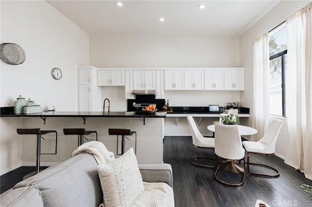 kitchen featuring a breakfast bar area, under cabinet range hood, dark wood finished floors, white cabinetry, and dark countertops