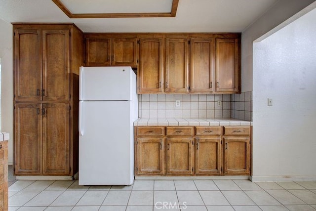 kitchen with freestanding refrigerator, light tile patterned floors, brown cabinetry, and tasteful backsplash