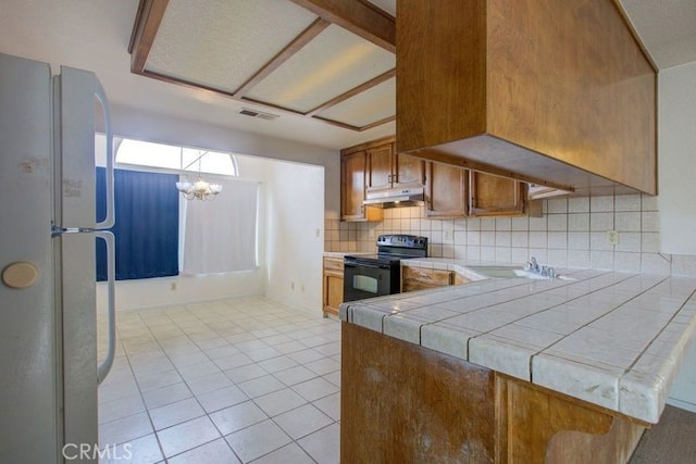 kitchen featuring under cabinet range hood, an inviting chandelier, black / electric stove, visible vents, and tile counters
