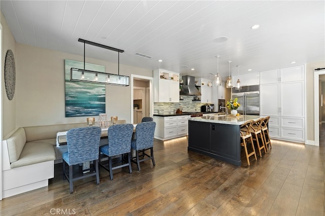 kitchen featuring stainless steel built in refrigerator, wall chimney range hood, white cabinets, dark wood finished floors, and an island with sink