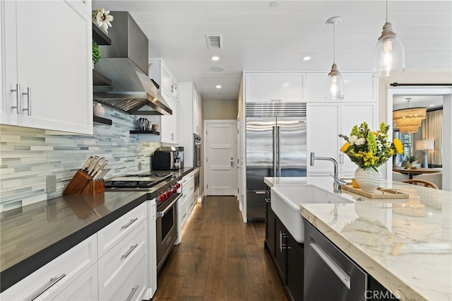 kitchen featuring high quality appliances, a sink, wall chimney range hood, dark stone countertops, and white cabinetry