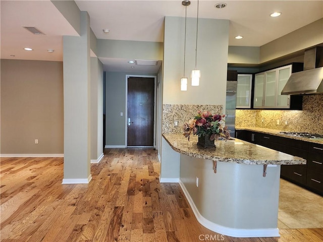kitchen with a kitchen breakfast bar, stainless steel gas stovetop, wall chimney exhaust hood, pendant lighting, and light stone countertops