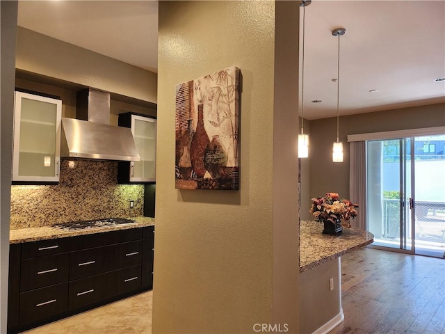 kitchen with glass insert cabinets, wall chimney range hood, tasteful backsplash, dark cabinetry, and stainless steel gas cooktop