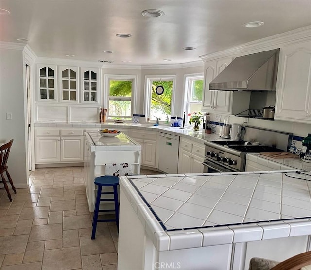 kitchen featuring wall chimney range hood, tile counters, stone tile flooring, white cabinets, and dishwasher