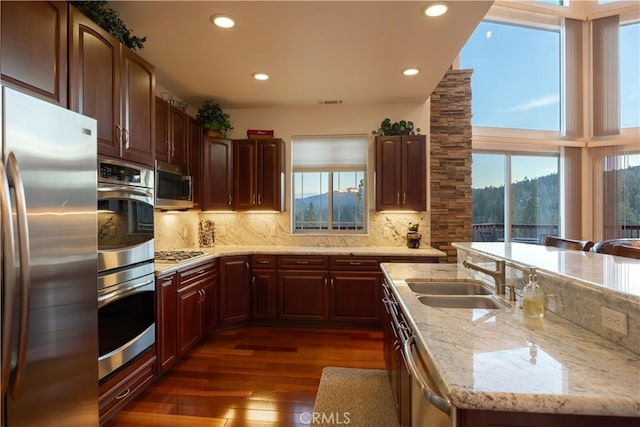 kitchen featuring light stone counters, a sink, backsplash, appliances with stainless steel finishes, and dark wood-style floors
