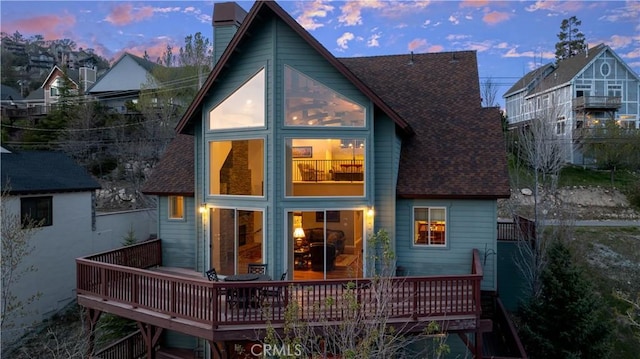 back of house at dusk featuring roof with shingles, a wooden deck, and a chimney
