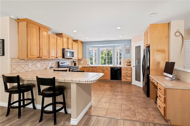 kitchen with black appliances, a peninsula, backsplash, a kitchen bar, and light brown cabinetry
