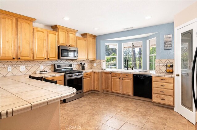 kitchen with stainless steel appliances, tile countertops, decorative backsplash, light brown cabinetry, and recessed lighting