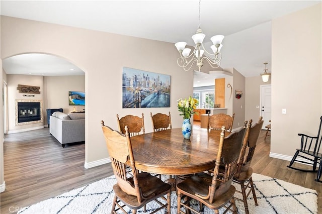 dining space featuring wood finished floors, arched walkways, baseboards, a chandelier, and a glass covered fireplace