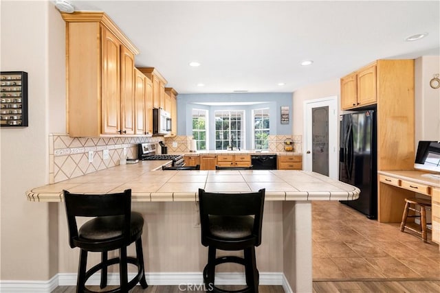 kitchen featuring black appliances, a peninsula, backsplash, light brown cabinetry, and a kitchen breakfast bar