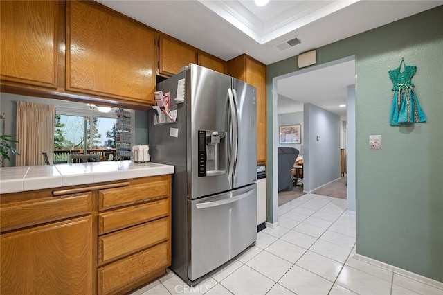 kitchen with light tile patterned floors, brown cabinetry, tile counters, stainless steel refrigerator with ice dispenser, and visible vents