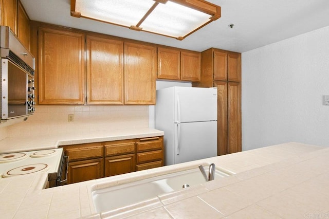 kitchen featuring stainless steel microwave, brown cabinetry, freestanding refrigerator, tile countertops, and backsplash