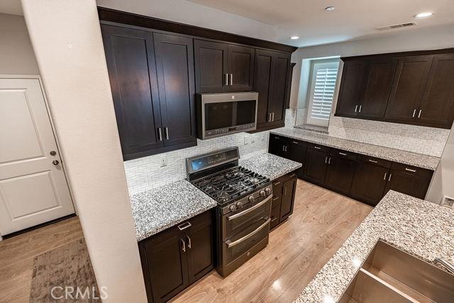 24189 Darmera Dr, Lake Elsinore, CA 92532 - kitchen featuring appliances with stainless steel finishes, light wood-type flooring, decorative backsplash, light stone counters, and dark brown cabinetry