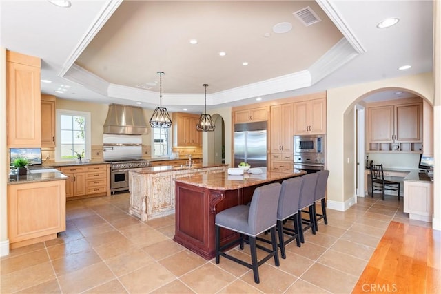 kitchen with premium range hood, built in appliances, a kitchen island with sink, and a raised ceiling