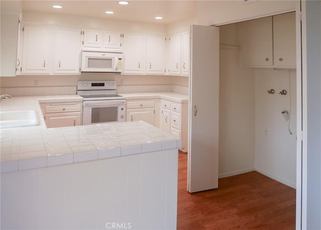 kitchen with a sink, a peninsula, white appliances, wood finished floors, and tile counters