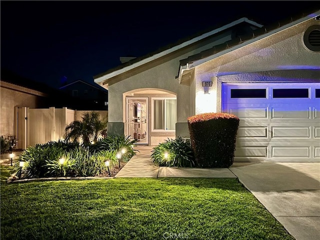 exterior entry at twilight featuring a lawn, stucco siding, a garage, and fence