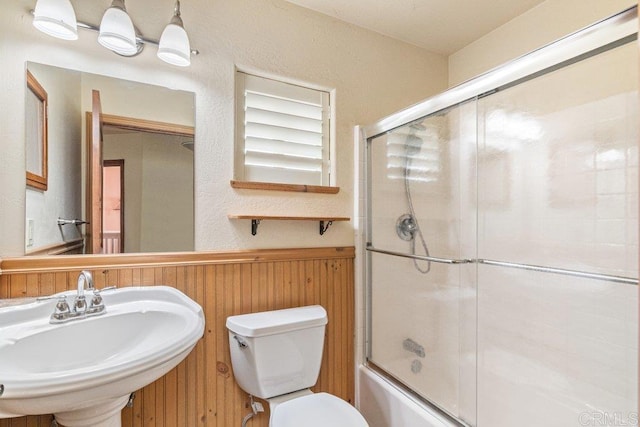 bathroom with toilet, shower / bath combination with glass door, a sink, wood walls, and wainscoting
