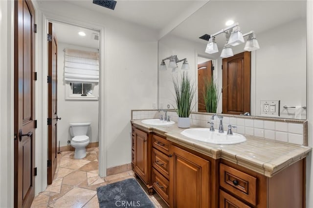 full bathroom featuring baseboards, double vanity, stone tile floors, toilet, and recessed lighting