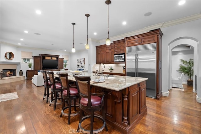 kitchen featuring built in appliances, arched walkways, dark wood-style floors, crown molding, and a warm lit fireplace