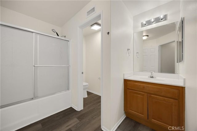 bathroom with toilet, wood finished floors, vanity, baseboards, and bath / shower combo with glass door