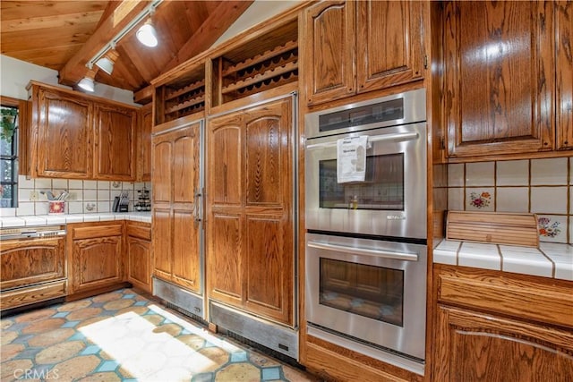 kitchen featuring stainless steel double oven, tile counters, tasteful backsplash, brown cabinets, and wood ceiling