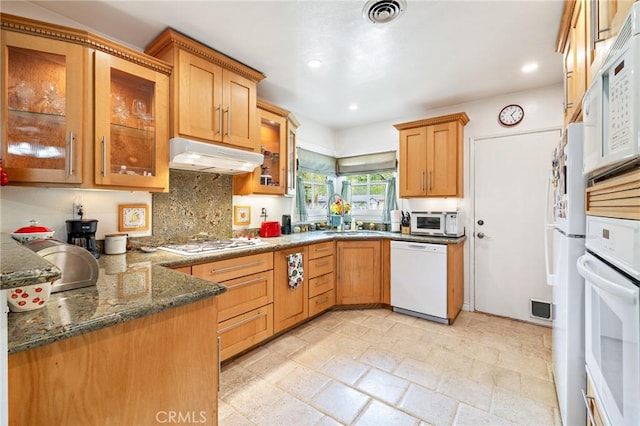 kitchen with white appliances, under cabinet range hood, dark stone counters, glass insert cabinets, and light stone finish floors