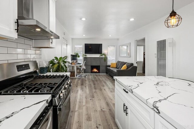 kitchen with stainless steel gas range oven, wall chimney exhaust hood, a heating unit, a tile fireplace, and light wood-type flooring