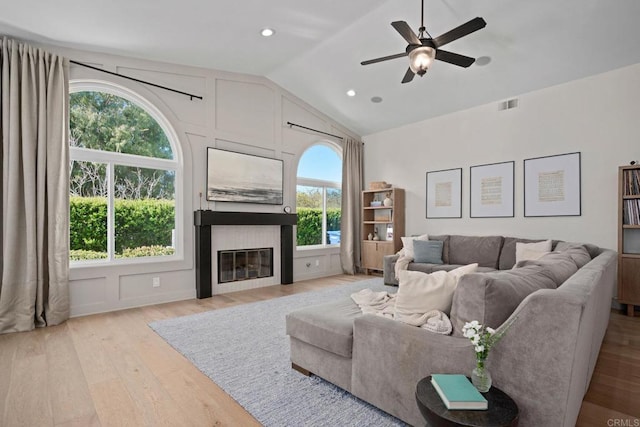 living area featuring lofted ceiling, light wood-style floors, a glass covered fireplace, and a ceiling fan