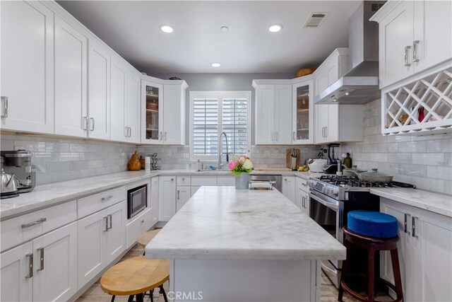 kitchen featuring wall chimney range hood, stainless steel appliances, a sink, a kitchen breakfast bar, and white cabinets