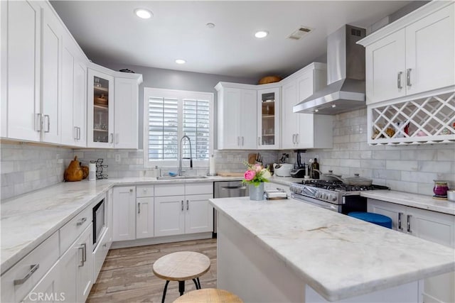 kitchen featuring wall chimney range hood, a sink, stainless steel range with gas cooktop, white cabinetry, and light wood-style floors