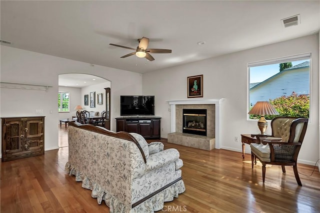 living room featuring a ceiling fan, arched walkways, wood finished floors, a tiled fireplace, and baseboards