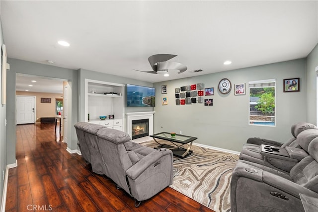 living area featuring a glass covered fireplace, a ceiling fan, dark wood finished floors, recessed lighting, and baseboards